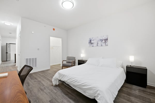 Bright studio bedroom with queen bed, gray armchair, desk, and modern ceiling lights