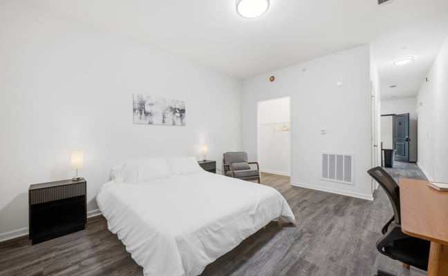 Spacious modern bedroom with queen bed, gray armchair, desk, and ensuite bathroom entrance